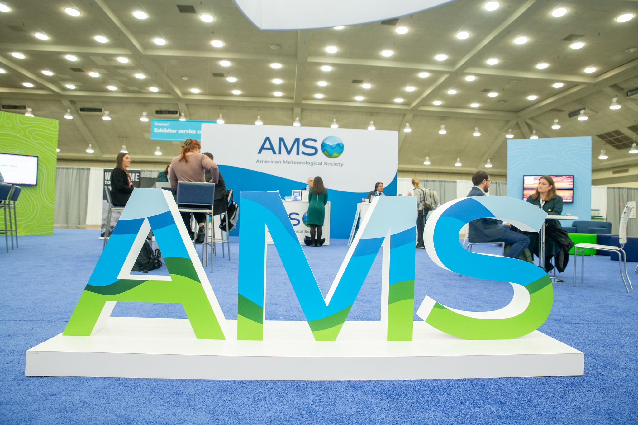 Large, colorful "AMS" letters prominently displayed on a blue carpeted exhibition floor. Attendees converse in a well-lit conference space.