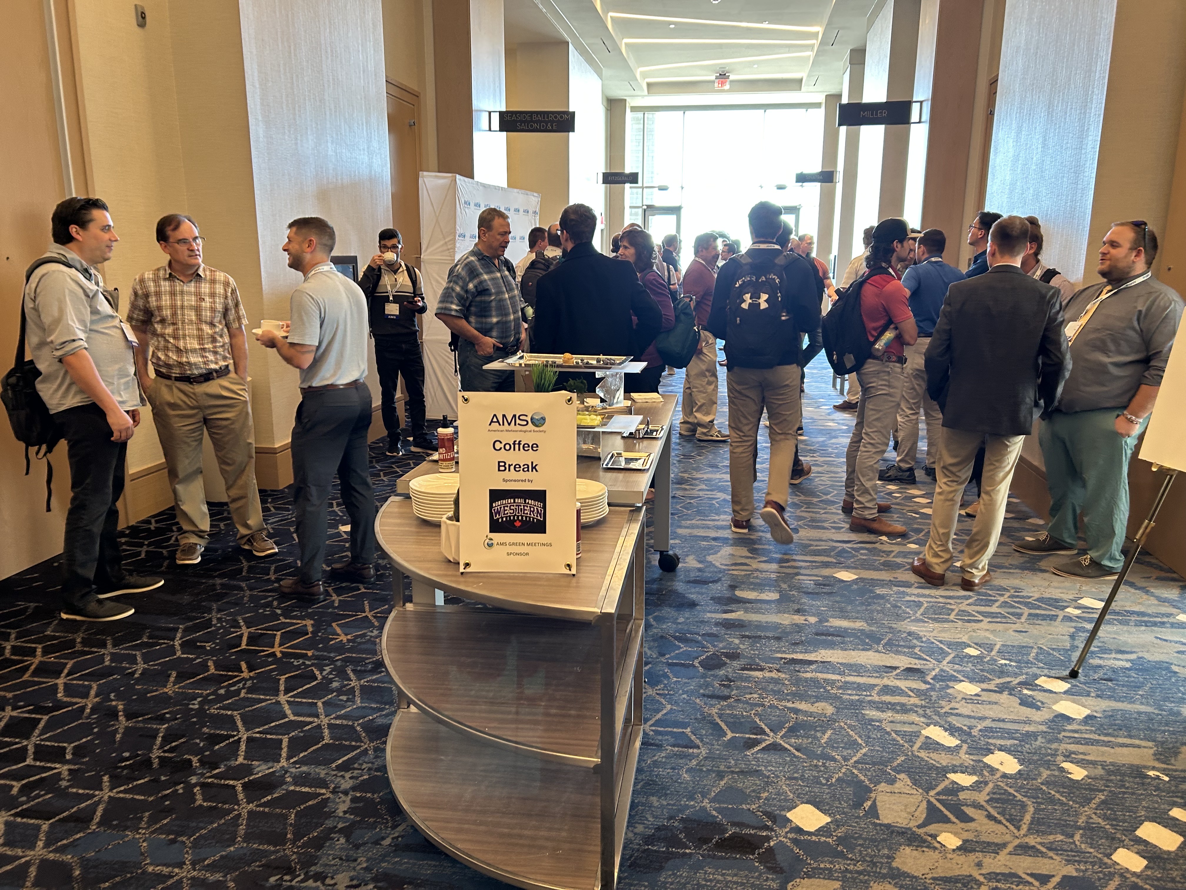 People gather in a conference hallway during a coffee break, chatting in groups. A sign on a table reads "AMS Coffee Break," creating a lively, networking atmosphere.