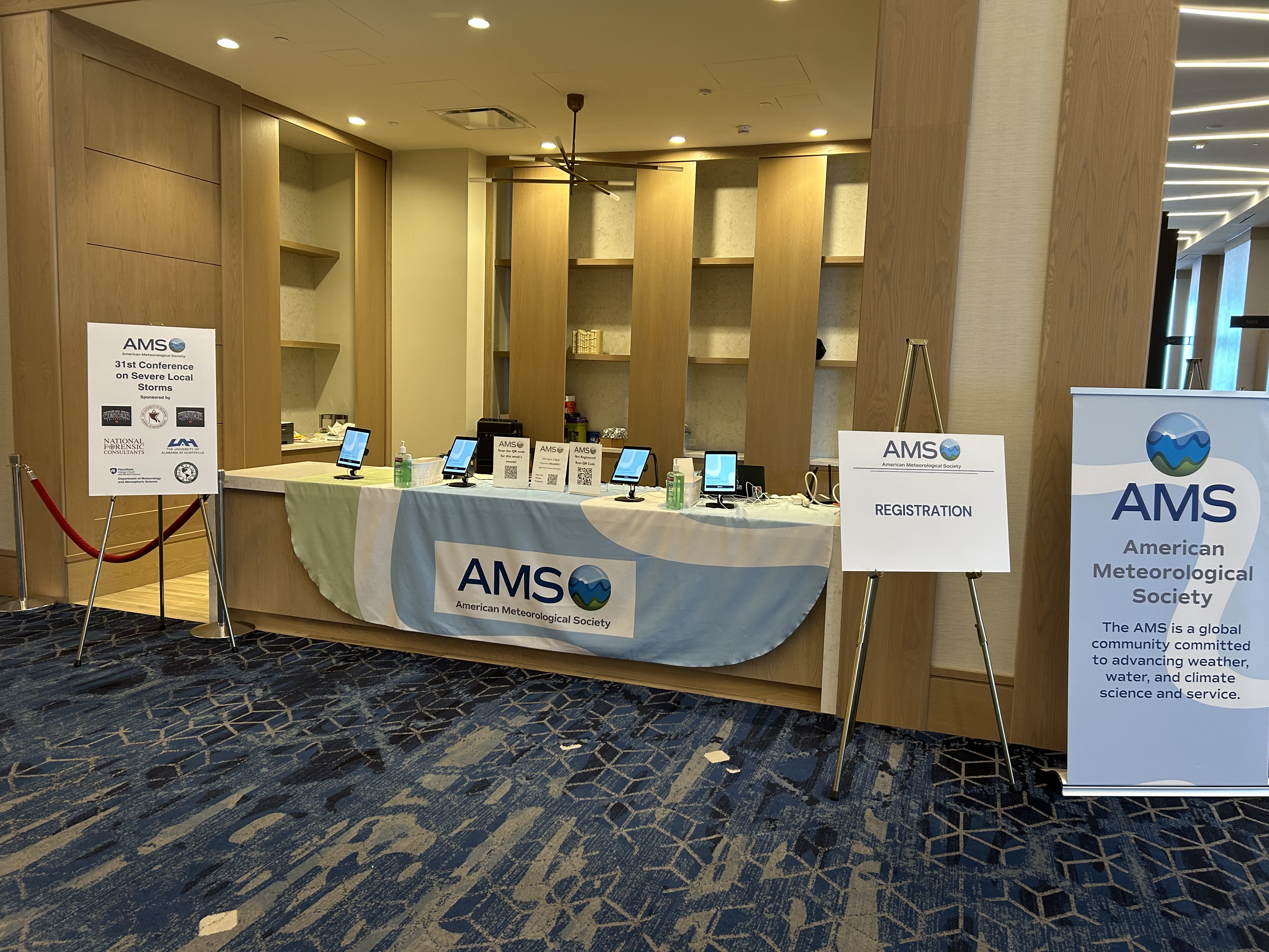 Lobby with AMS registration desk for a meteorological conference. Blue carpet, signs, and tablets on a table covered with a logo-themed cloth.