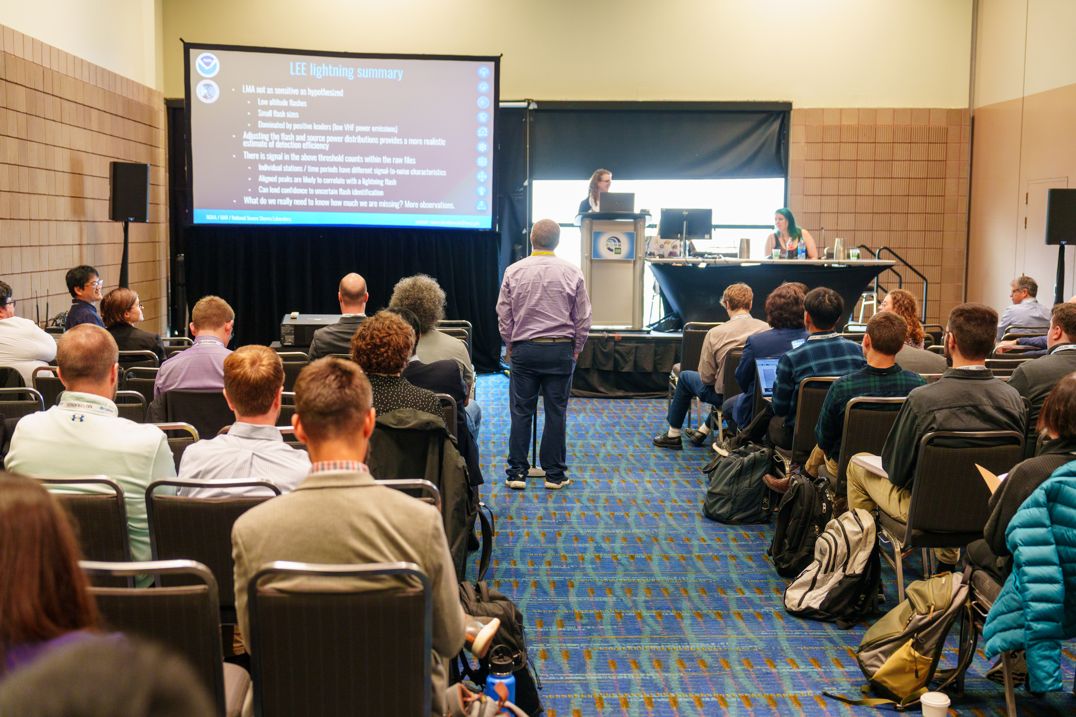 A conference room filled with seated attendees facing away from the camera and towards a speaker at a podium. An attendee is standing in the aisle near the stage to ask the presenter at the podium a question. A presentation screen on the upper left side of the image displays slides. 